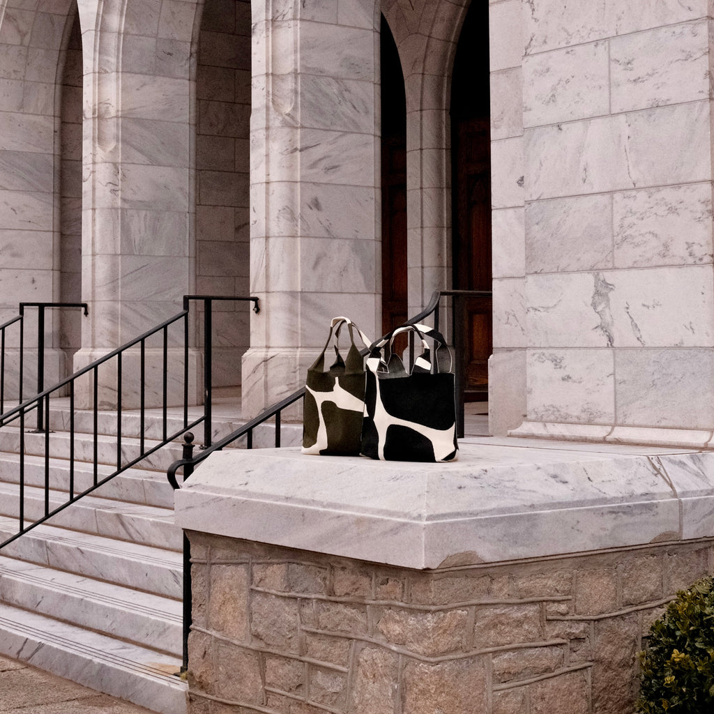 MOOdy Black Signature Tote bag displayed alongside a green and white patterned tote outside an elegant building.