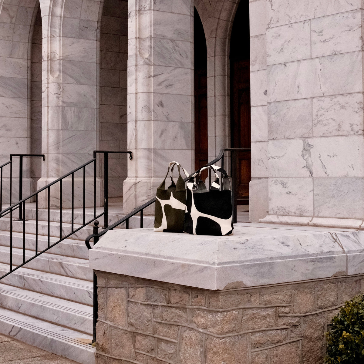 MOOdy Black Signature Tote bag displayed alongside a green and white patterned tote outside an elegant building.